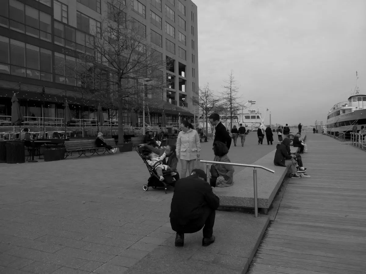 People on a waterfront promenade with buildings and a boat.